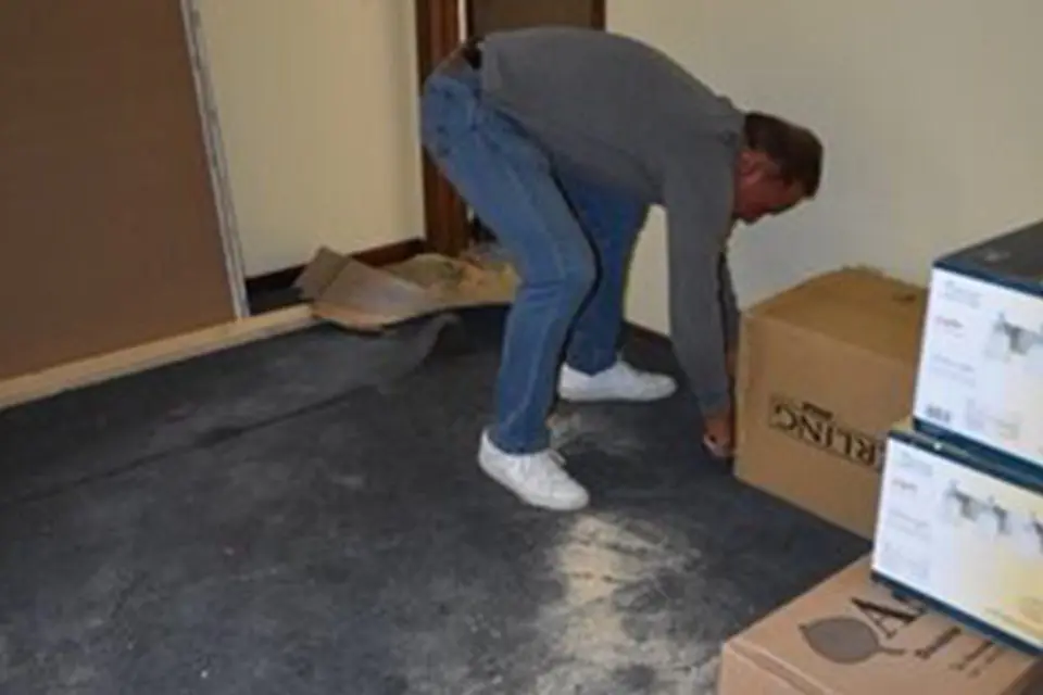A person in jeans and a gray shirt bends down to lift a cardboard box in a sparsely furnished room with a bare floor.
