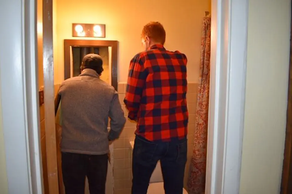 Two men stand side by side facing a toilet in a small bathroom, seen from behind through an open doorway.