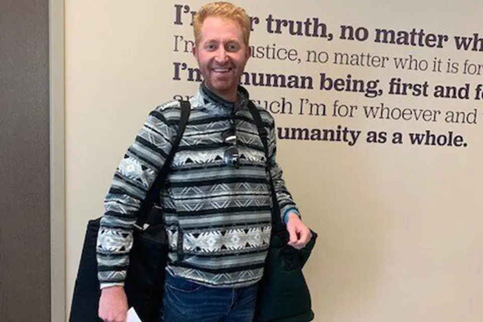 A man with red hair stands indoors, wearing a patterned sweater and holding a large black bag; text with inspirational quotes is visible on the wall behind him.