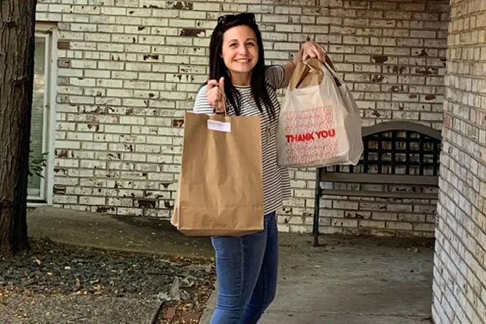 A woman stands outside a brick building, holding two shopping bags and smiling at the camera.