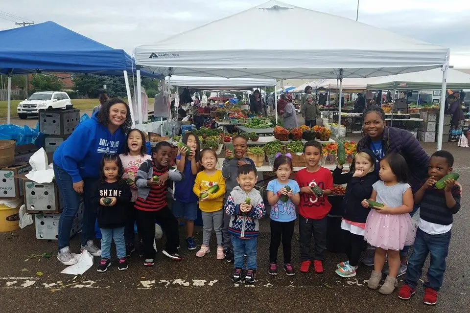 A group of children and two adults stand under tents at an outdoor market, each child holding vegetables and smiling at the camera.