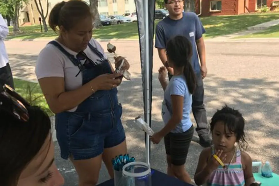 A woman checks her phone while standing with three children under a canopy outdoors; one child eats a treat and another holds a yellow item.