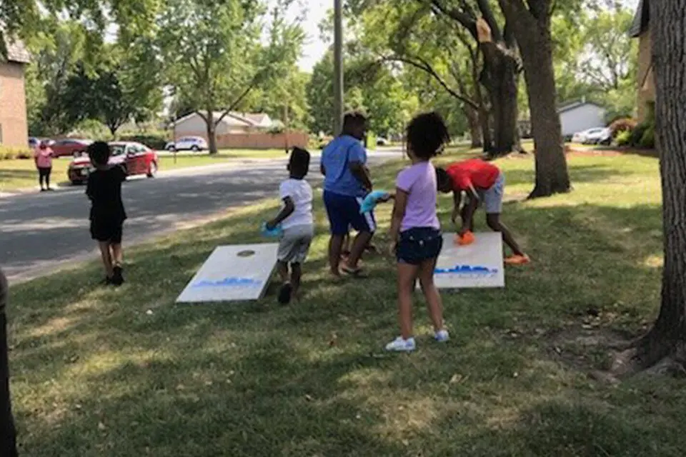 Six children are playing cornhole on a grassy area near a street on a sunny day, with trees and houses in the background.