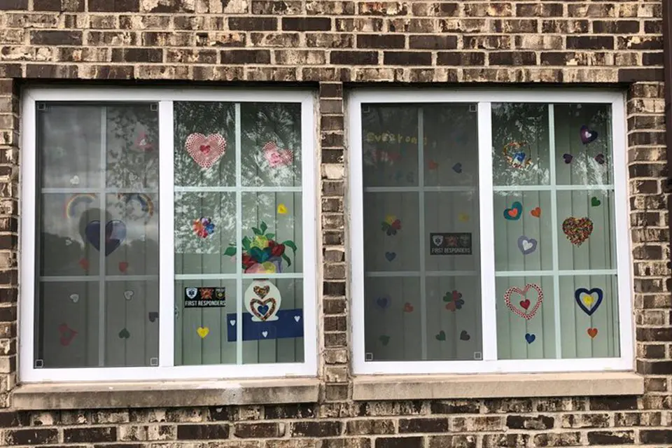 Two windows on a brick building are decorated with colorful paper hearts, shapes, and encouraging signs taped to the glass.