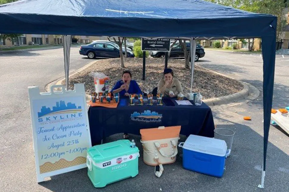 Two people sit under a blue canopy at a table with coolers, drinks, and containers, next to a sign promoting a tenant appreciation ice cream party event.