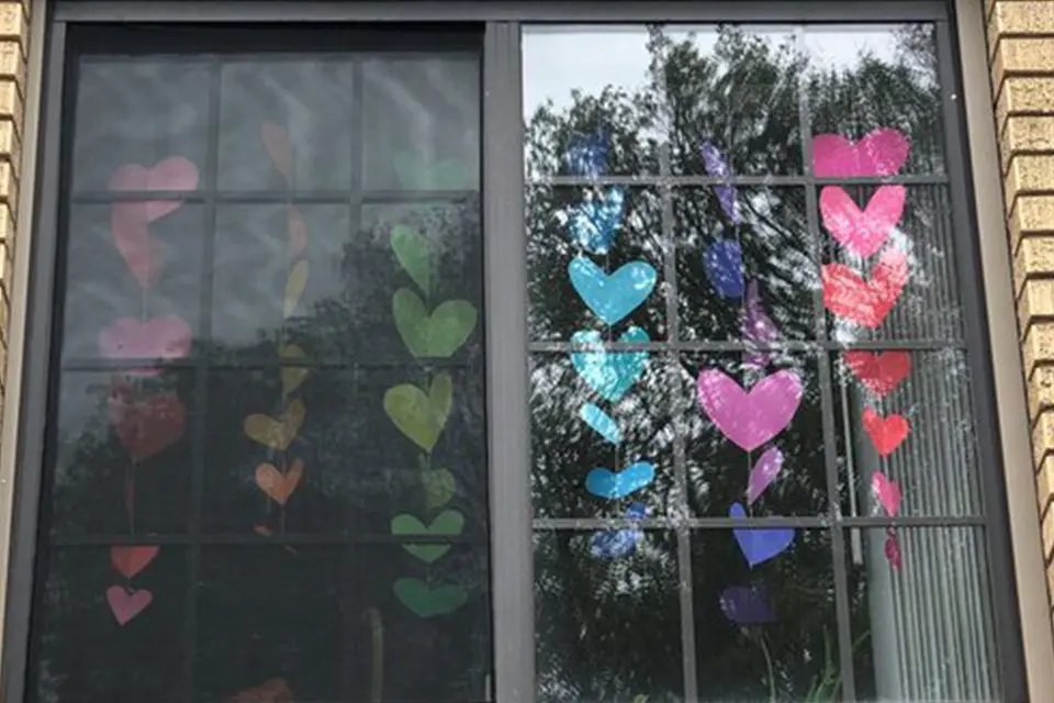 A window decorated with colorful paper hearts arranged in vertical rows, with trees and sky reflected in the glass.
