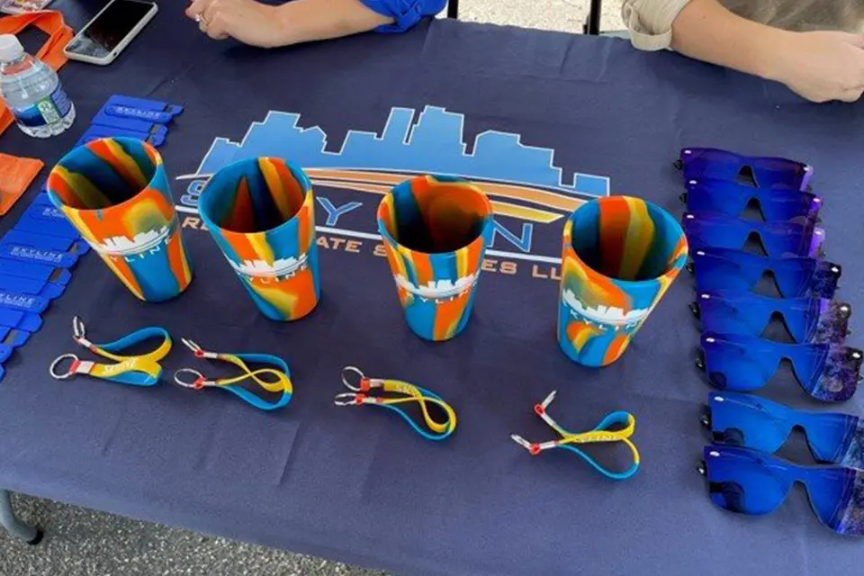 Table with promotional items including three colorful cups, blue sunglasses, branded keychains, and business card holders on a blue tablecloth with a city skyline logo.