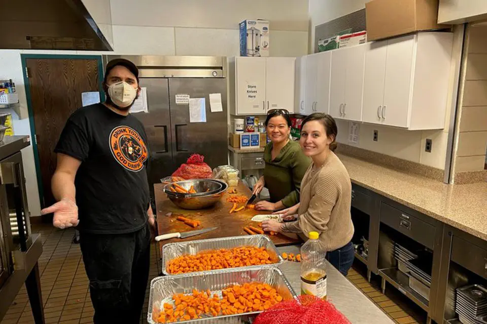 Three people in a commercial kitchen, two cutting carrots at a table, and one standing nearby wearing a mask. Trays of chopped carrots are on the counter.