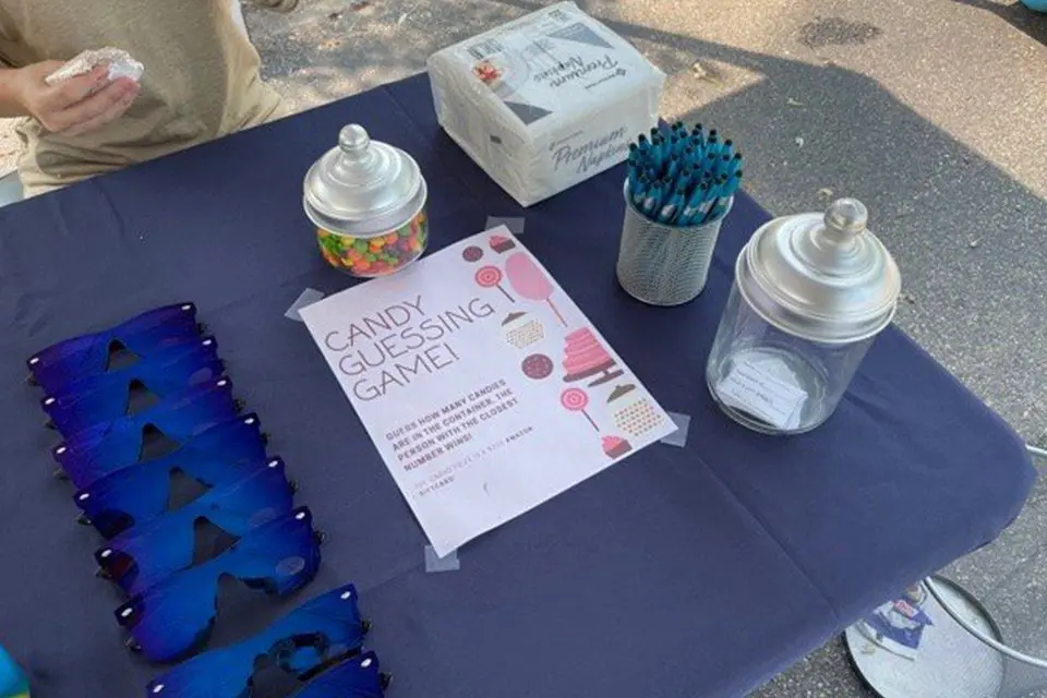 A table with a "Candy Guessing Game" sign, jars of candy and paper slips, blue sunglasses, a box of napkins, and a cup of blue pens on a dark tablecloth.