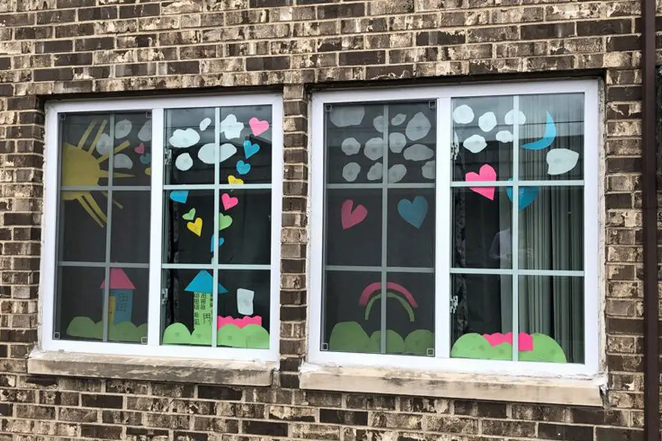 Two windows with colorful paper cutouts of hearts, clouds, a sun, a crescent moon, a rainbow, and houses displayed on the glass of a brick building.