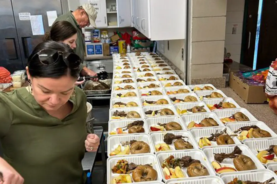 Three people prepare and pack numerous meal trays with bagels, meat, and fruit on a kitchen counter.