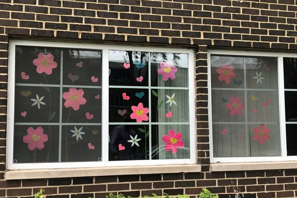 A brick building window is decorated with large pink paper flowers and colorful paper hearts.