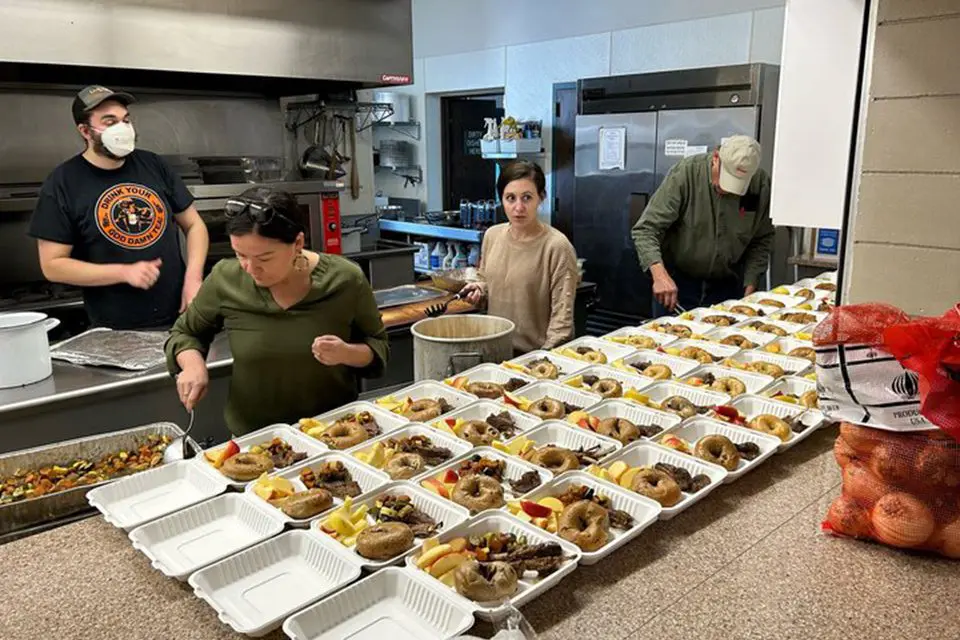 Four people prepare and pack food into takeout containers in a commercial kitchen, with rows of containers filled with meals lined up on the counter.