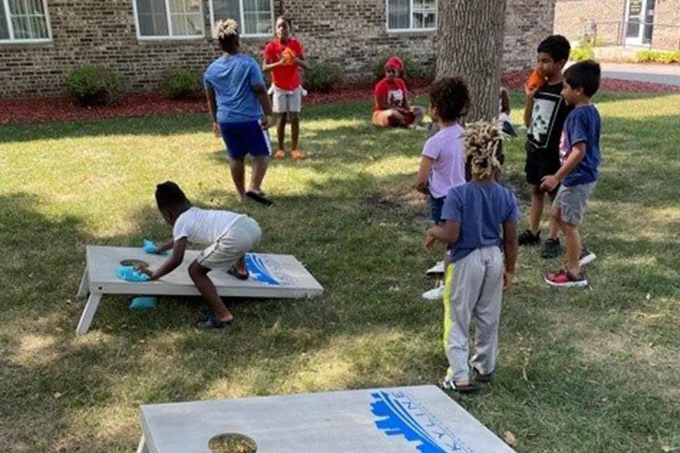 Several children play a bean bag toss game on a grassy area outside, with two adults sitting nearby against a tree and a brick building in the background.