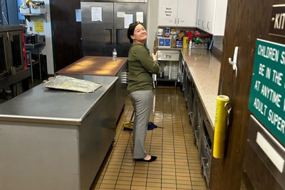 A woman stands and smiles at the camera while working in a commercial kitchen, with cleaning supplies and utensils visible on the counter.