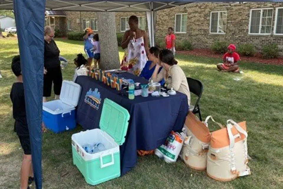 People gather under a canopy at an outdoor event with a table displaying drinks and snacks, coolers, and bags on the grass. A brick building is visible in the background.