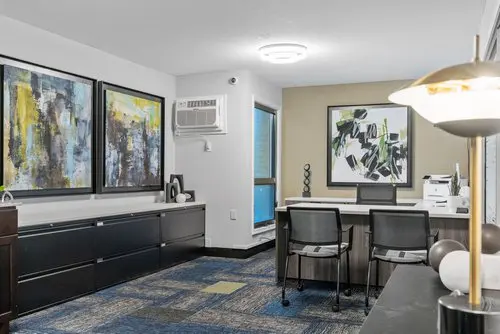 Modern office space with two chairs at a desk, abstract artwork on the walls, filing cabinets, and a wall-mounted air conditioner.