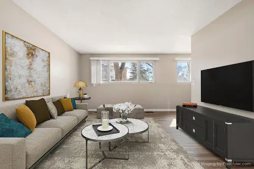 A modern living room with a beige sofa, colorful pillows, a coffee table, large wall art, a TV on a black console, and windows letting in natural light.