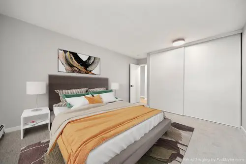 Modern bedroom with a gray upholstered bed, orange blanket, bedside tables with lamps, abstract wall art, and large white sliding closet doors.