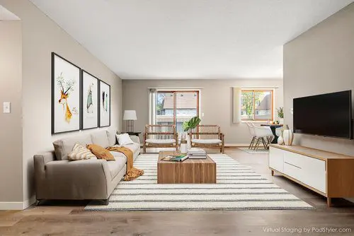 A modern living room with a gray sofa, striped rug, coffee table, wall art, TV on a white stand, and a dining area with windows in the background.