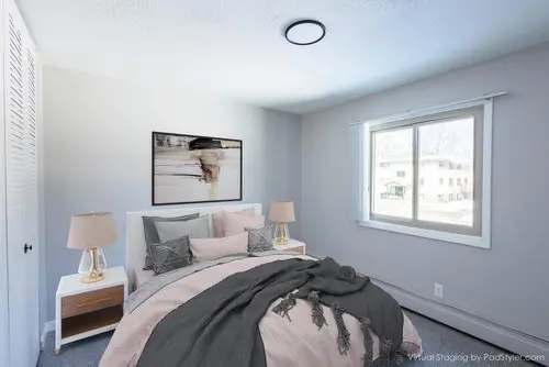A bedroom with a neatly made bed, two bedside tables with lamps, a large window, and a framed abstract artwork on the wall.