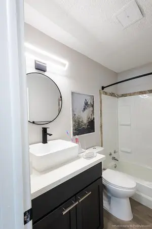 Modern bathroom with a round mirror, rectangular sink, black faucet, and dark vanity; bathtub with white shower curtain and wall art above the toilet.