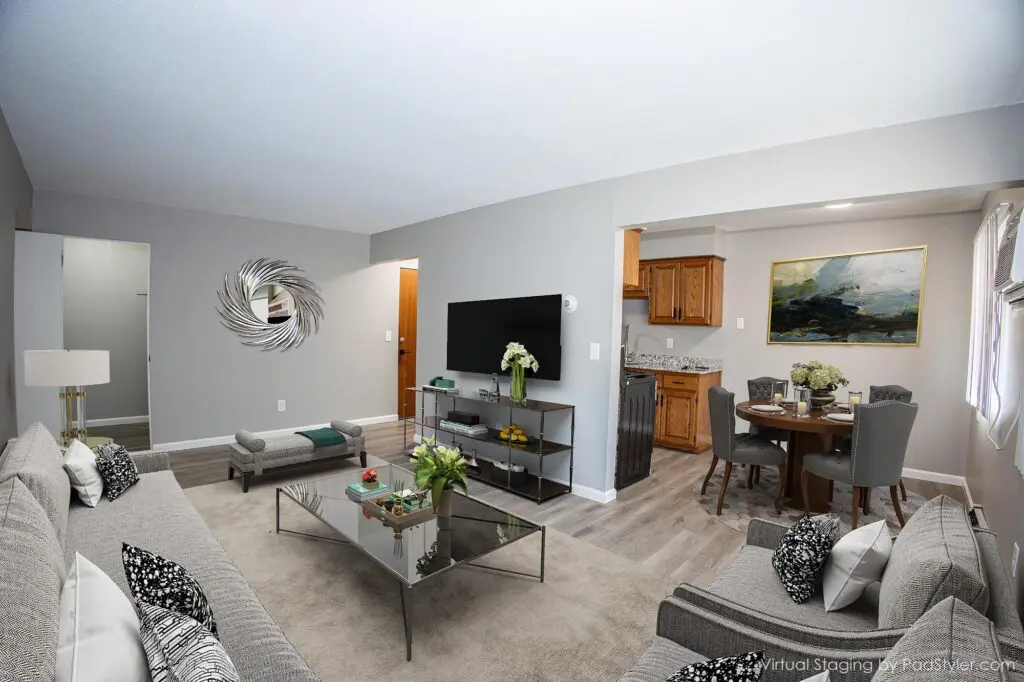 A modern, open-concept living and dining area with neutral tones, gray furniture, a wall-mounted TV, and a kitchen with wooden cabinets in the background.