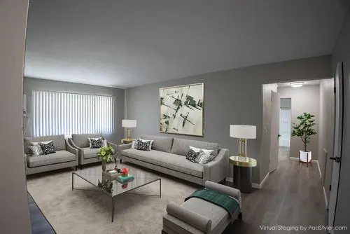 A modern living room with gray sofas, a glass coffee table, armchairs, and a large abstract painting on the wall. The room has wood flooring and a large window with blinds.