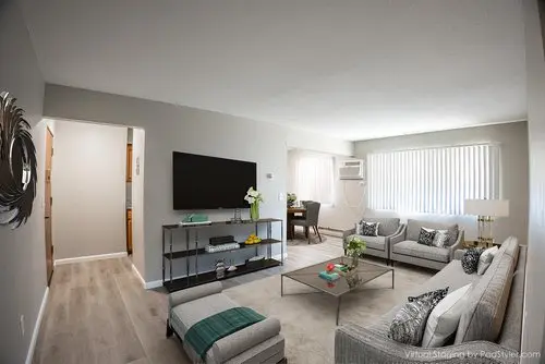 Modern living room with a wall-mounted TV, glass coffee table, gray sofas, decorative pillows, and large window with vertical blinds. Dining area visible in the background.