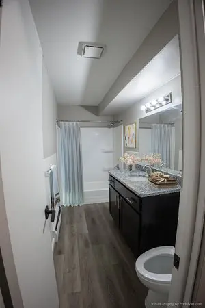 A modern bathroom with a shower and curtain, dark vanity with double sinks, lighted mirror, and toilet on a wood-style floor.