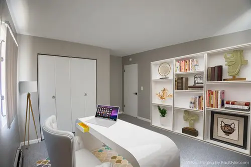 Modern home office with a white desk, laptop, white chair, and a large bookshelf filled with books and decorative items against a gray wall.