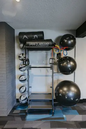 A metal gym rack holds black exercise balls, kettlebells, resistance bands, and two rolled-up exercise mats in a corner of a fitness room.
