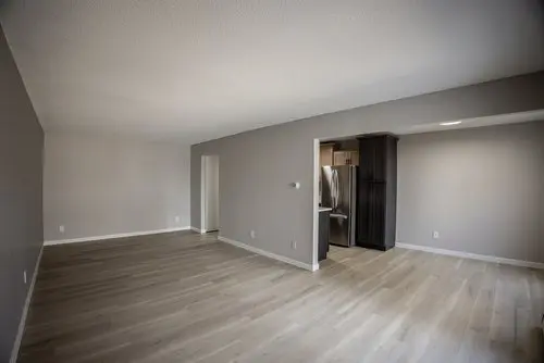 Empty room with light-colored wood flooring, gray walls, and an open doorway leading to a modern kitchen with stainless steel refrigerator.