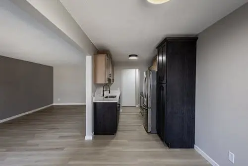 A modern, unfurnished kitchen with stainless steel appliances, dark cabinets, light wood flooring, and an open layout leading to a spacious living area.