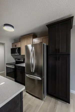 Modern kitchen with stainless steel fridge, black and wood cabinets, electric stove, and light-colored countertops and flooring.