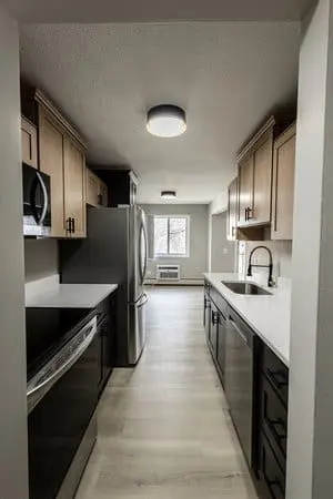 Modern galley kitchen with stainless steel appliances, light wood cabinets, white countertops, and a window at the far end providing natural light.