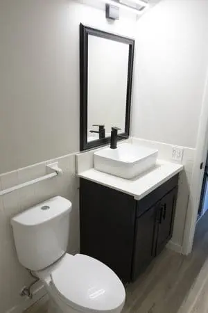 A small bathroom features a white toilet, a dark vanity with a white vessel sink, a black faucet, a rectangular mirror, and light wood flooring.