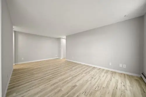 Empty room with light gray walls, light wood flooring, and baseboard trim; open layout with a narrow hallway visible in the back.