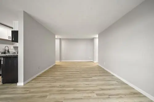 Empty room with light wood flooring, gray walls, and white trim; partial kitchen area with dark cabinets visible on the left side.