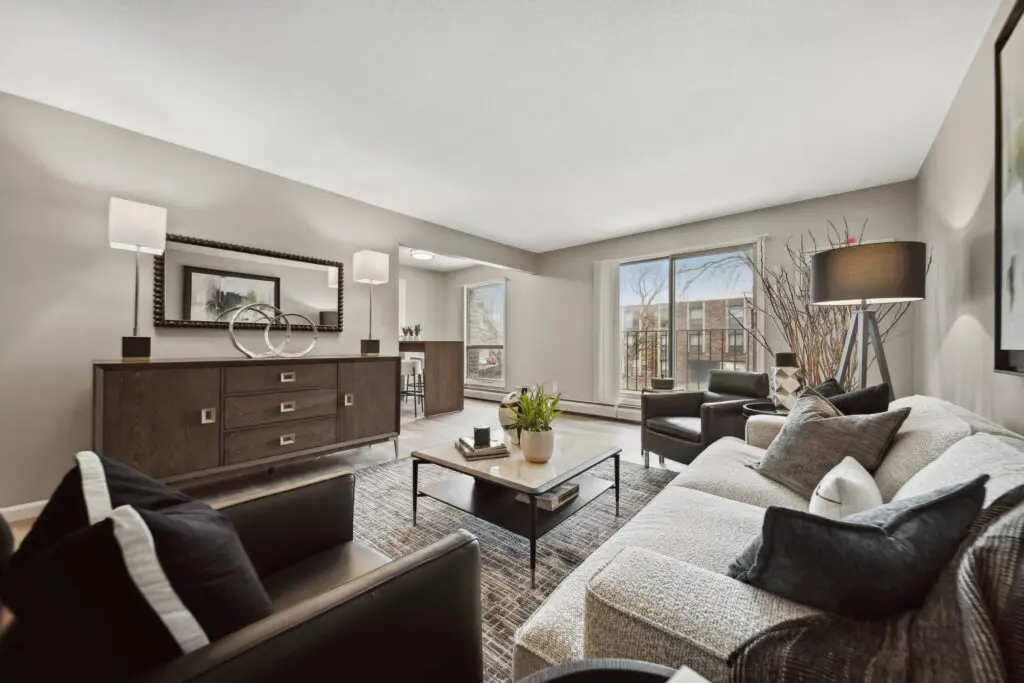 A modern living room with neutral tones features a sofa, two armchairs, a coffee table, sideboard, lamps, and large windows letting in natural light.