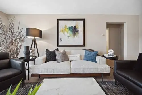 Modern living room with a white sofa, assorted cushions, two black armchairs, a floor lamp, abstract wall art, and decorative items on side tables.