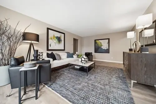 Modern living room with neutral tones, abstract wall art, a white sofa, two black chairs, a patterned rug, side tables, and a dark wood console table with lamps and decor.