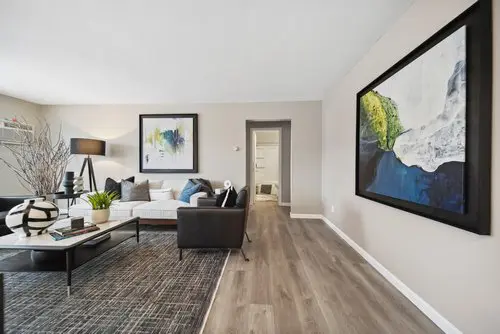 Modern living room with neutral walls, wood flooring, abstract artwork, black and white furniture, and a coffee table with decorative items. Doorway leads to another room.