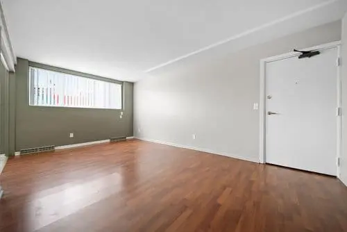 Empty room with light gray walls, wooden floor, a large window with blinds, and a white door.