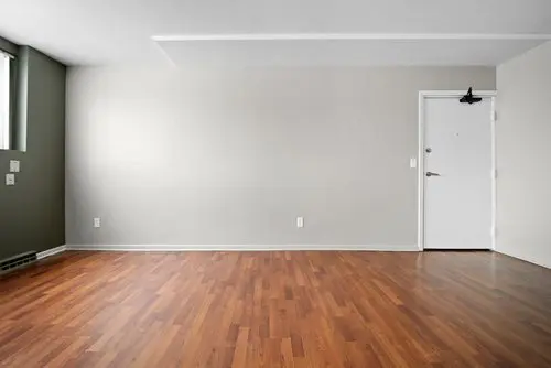 An empty room with light gray walls, a white door, wooden floors, and a window on the left letting in natural light.