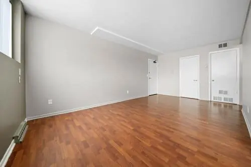 Empty room with light gray walls, white ceiling, and wooden floor; two white doors and a window on the left side.