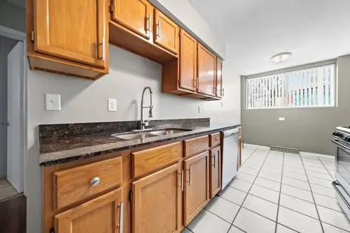 Kitchen with wooden cabinets, stainless steel sink, dark countertops, tile floor, and large window with vertical blinds letting in natural light.