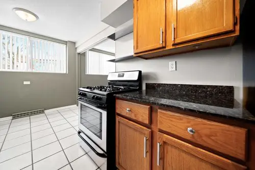 Kitchen with a stainless steel gas stove, black countertop, wooden cabinets, white tile floor, and large windows with vertical blinds.