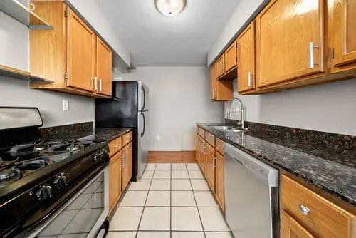 Galley kitchen with wood cabinets, black granite countertops, stainless steel appliances, gas stove, tiled floor, and neutral wall color.
