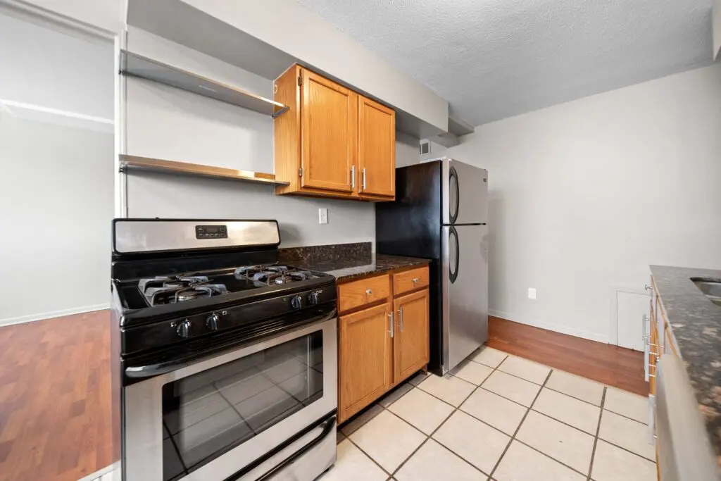 Modern kitchen with a stainless steel gas stove, wooden cabinets, black granite countertops, stainless steel refrigerator, and tile flooring.